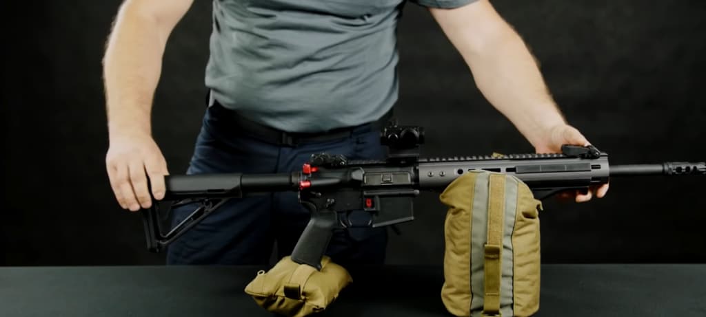 A person holds a modern rifle with attachments against a black background, with a tan bag below