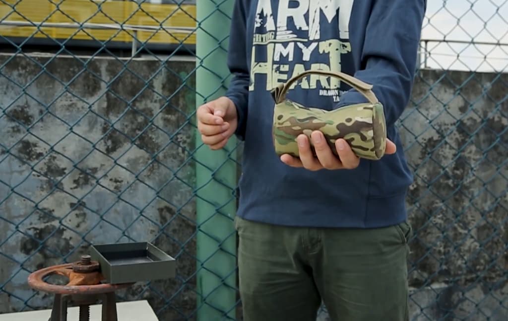 Person in blue shirt holds camo pouch near fence and rusty valve