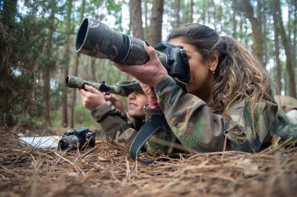 Two individuals; one capturing photos in a forest, the other observing through binoculars